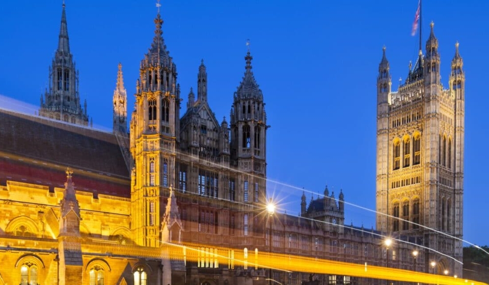 Long exposure night shot of the Houses of Parliament in London with blue sky and a street in the foreground.